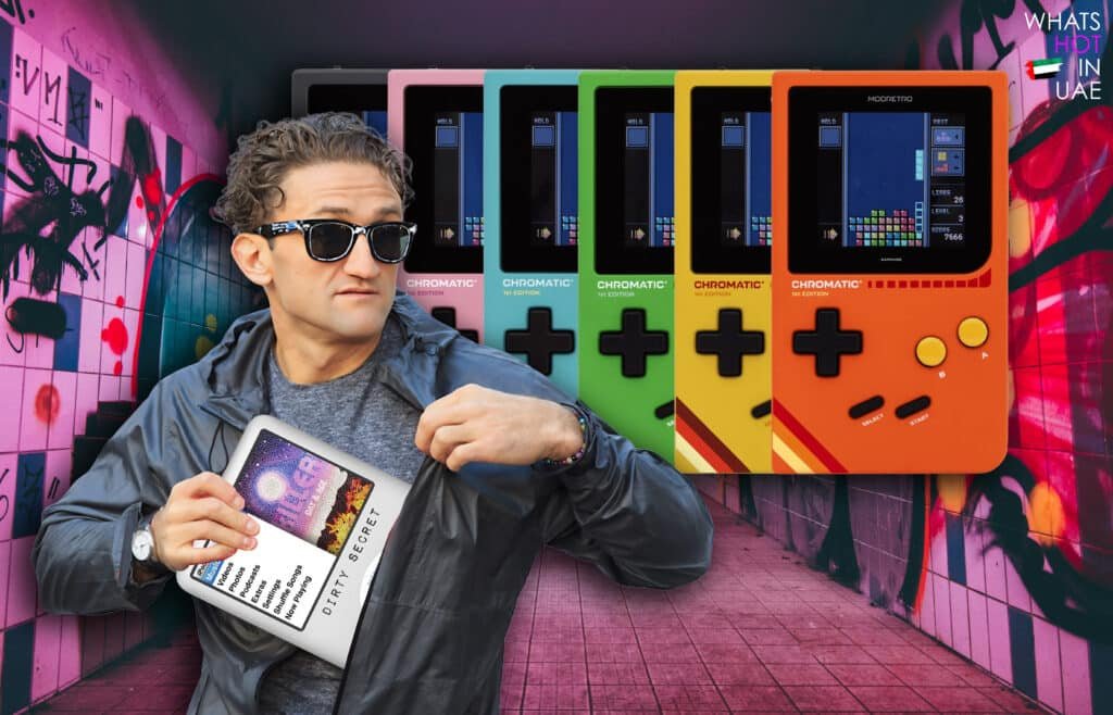 Casey Neistat in sunglasses and a grey jacket is pulling an iPod from his pocket, revealing a screen displaying text. Behind him are several colorful retromod gaming consoles labeled "CHROMATIC 1st EDITION" in various colors. The background features a graffiti-covered wall with pink tones.
