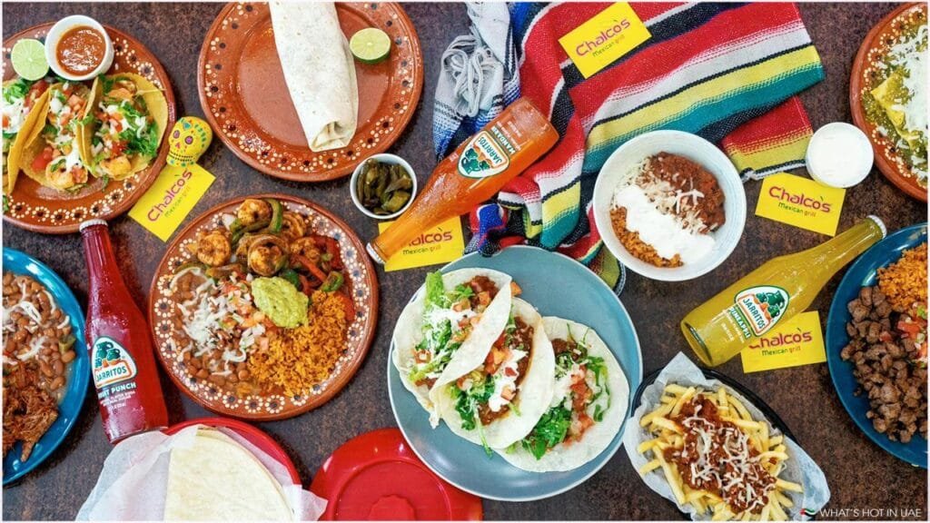 An assortment of Mexican dishes including tacos, a burrito, nachos, rice, and beans, displayed on colorful plates with bottles of Jarritos soda and restaurant branding visible.