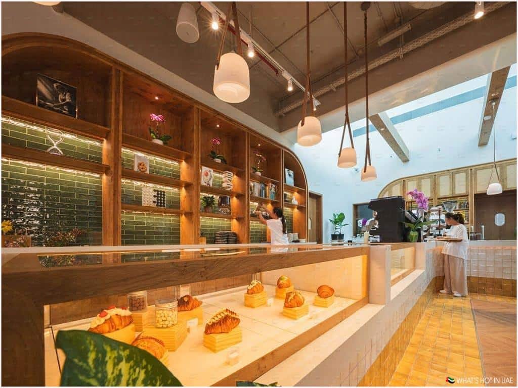 A modern café interior with a warm, inviting atmosphere. The space features a wooden counter displaying various pastries and jars. The back wall has shelves lined with decorative items and green tiled accents. Two people are attending to tasks behind the counter, which is illuminated by hanging pendant lights. Potted plants add a touch of greenery to the setting.