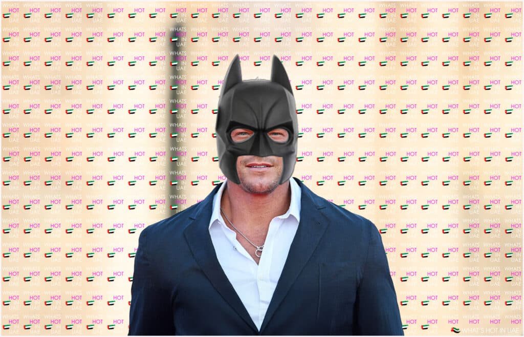 A person wearing a Batman mask is standing in front of a patterned background that reads "WHAT'S HOT IN UAE" with small flag icons.