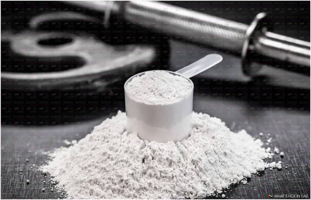 A scoop filled with white Creatine powder on a pile of the same powder, with a barbell and weight plate in the background.