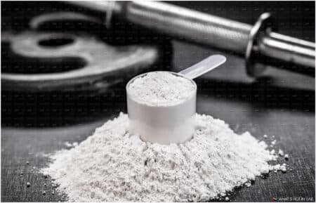 A scoop filled with white Creatine powder on a pile of the same powder, with a barbell and weight plate in the background.