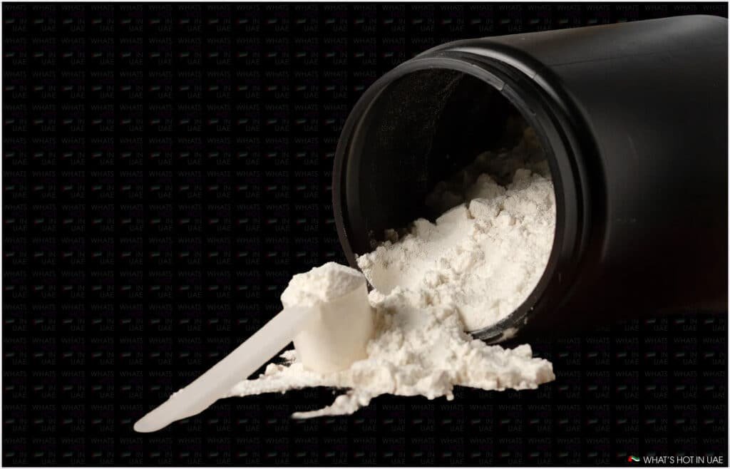A black container of white powder tipped over, spilling its contents onto a surface, with a small plastic scoop filled with the powder lying in front.