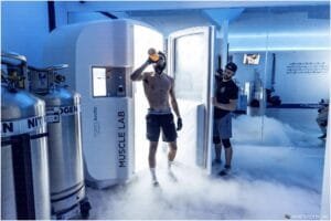 A shirtless man drinking from an orange bottle steps out of a cryotherapy chamber labeled 'Muscle Lab,' with misty fog covering the floor. Another man, wearing a cap and shorts, stands nearby holding the door open. The chamber is situated in a modern gym environment with reflective surfaces.