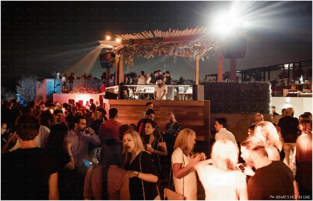 A crowded rooftop bar at night, filled with people socializing and a DJ performing in the background, under a well-lit canopy with hanging plants.