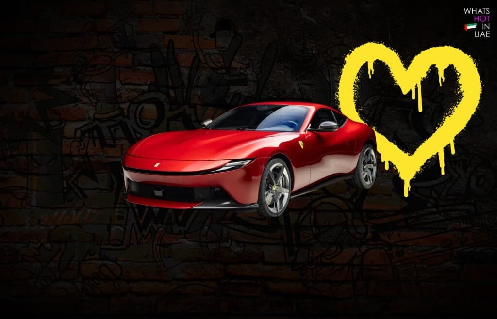 A red Ferrari sports car is prominently displayed against a dark, graffiti-covered wall, with a large yellow heart shape splattered nearby.