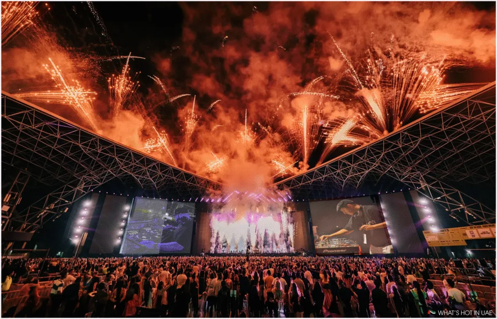A large crowd at an indoor music concert with a DJ performance by Smokingroove, surrounded by dramatic fireworks and lighting effects under a massive arched roof as a part of UAE Nightlife.