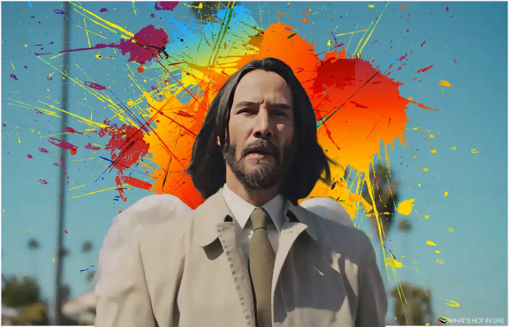 A person with long, dark hair and a beard is wearing a beige trench coat and tie, set against a vivid, abstract background with splashes of red, yellow, and blue paint on a bright blue sky.