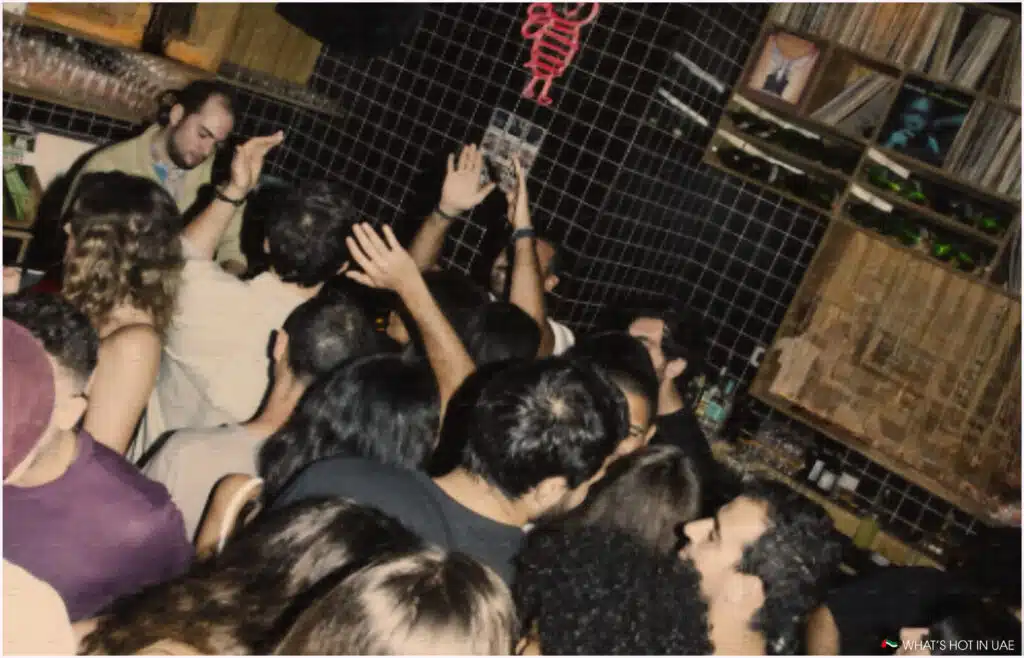 Crowded interior of a nightclub with people dancing and raising their hands, surrounded by dark tiled walls and shelves filled with bottles and records.