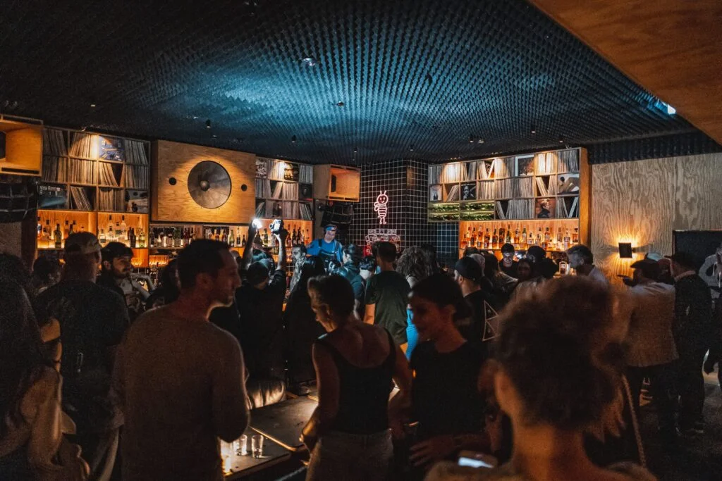 A crowded bar scene with people socializing, featuring shelves filled with liquor bottles and vinyl records, illuminated by warm lighting and a blue spotlight on the crowd.