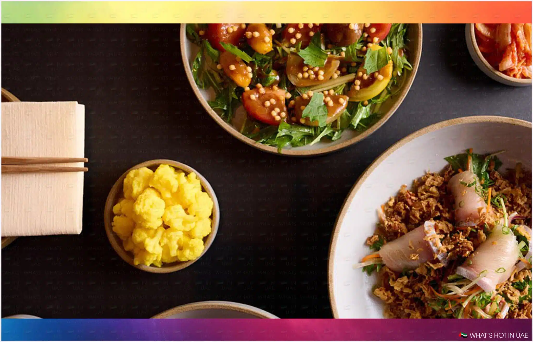 A table set with a variety of dishes including a salad with cherry tomatoes and greens, a bowl of yellow cauliflower pieces, and a plate of finely chopped vegetables with sliced radish and herbs. Wooden chopsticks rest on a napkin to the left.