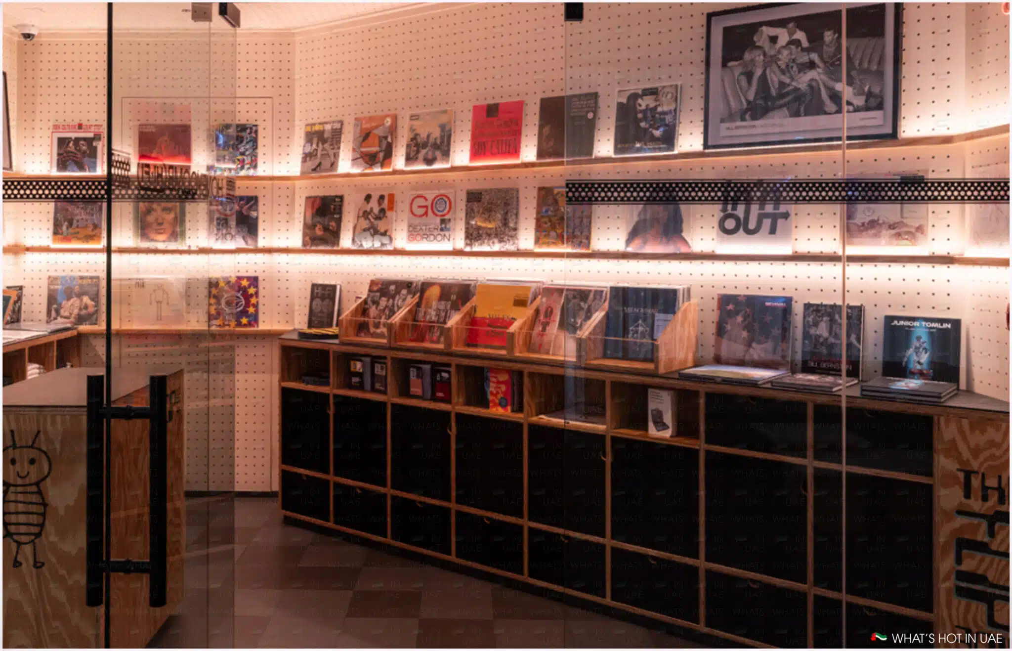 A record store display with wooden shelves and pegboard walls, showcasing various vinyl records and music-related items, lit by warm overhead lights. The store also features a wooden counter with a cartoon character drawing on the front.