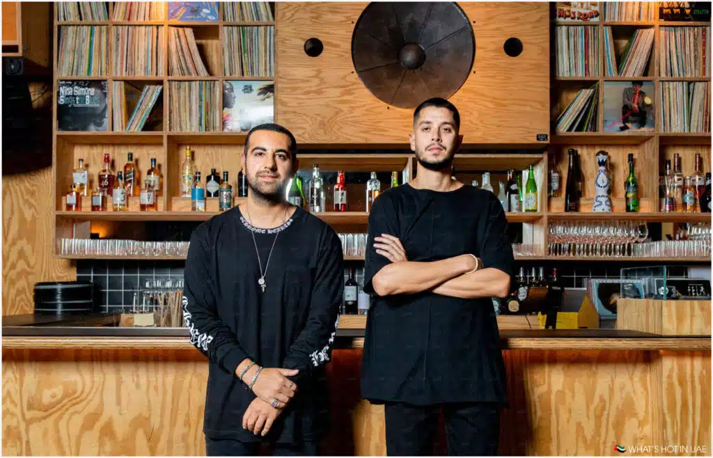 Two men standing in front of a bar, with shelves behind them displaying liquor bottles and vinyl records, in a stylish wooden interior.