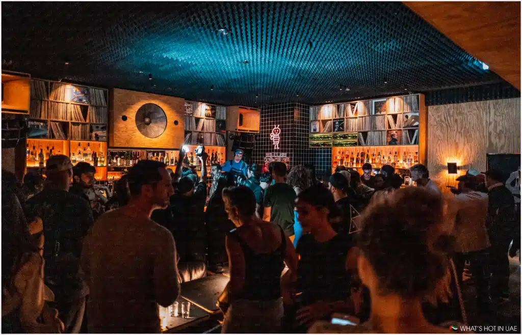 A crowded bar with people socializing, dim lighting, and shelves of records behind the bar area.