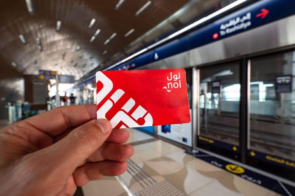 A hand holding a red Nol card in front of a platform in a metro station with signs in both Arabic and English.