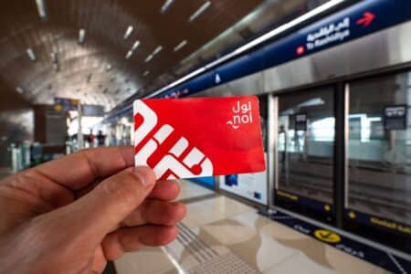 A hand holding a red Nol card in front of a platform in a metro station with signs in both Arabic and English.