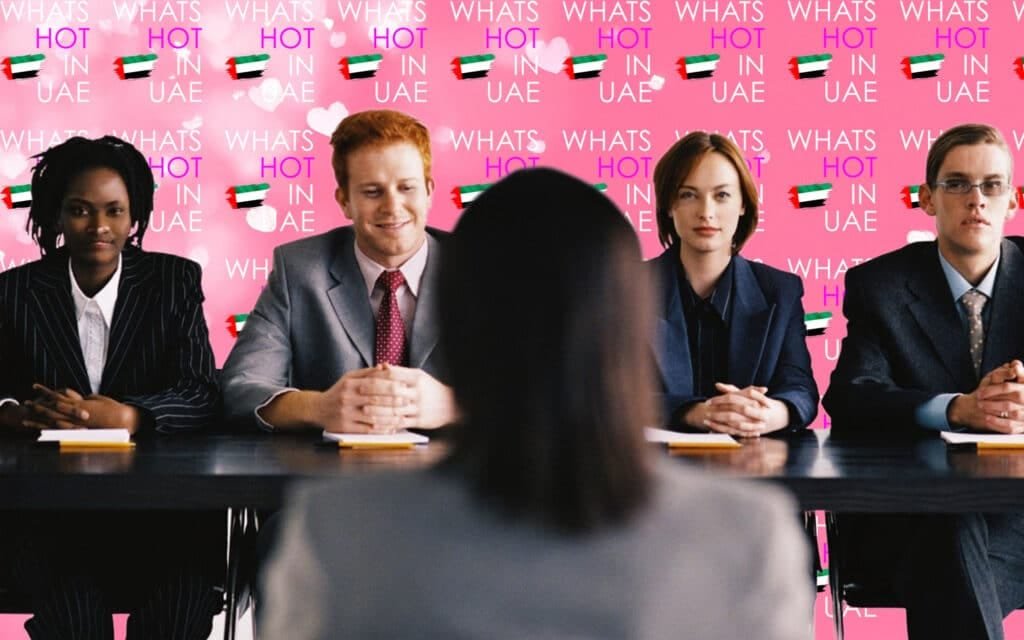 Four people sitting at a table dressed in business attire, with a pink background displaying repeated text 'WHATS HOT IN UAE' and UAE flags.