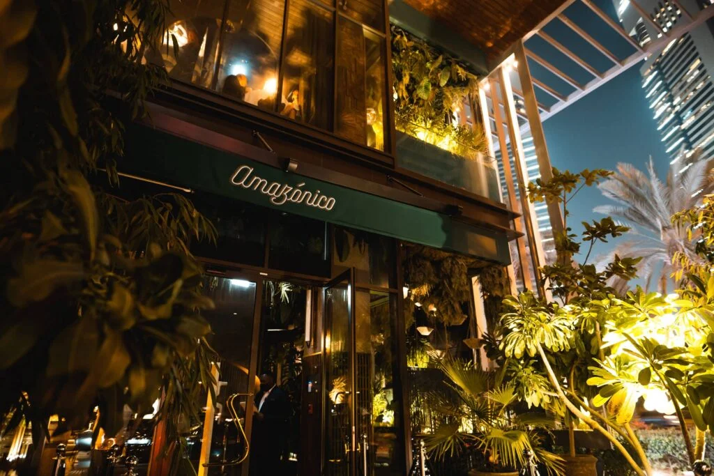 The entrance of a restaurant named "Amazonico" featuring lush greenery and modern lighting, with a doorman in front and a backdrop of urban buildings in the evening sky.