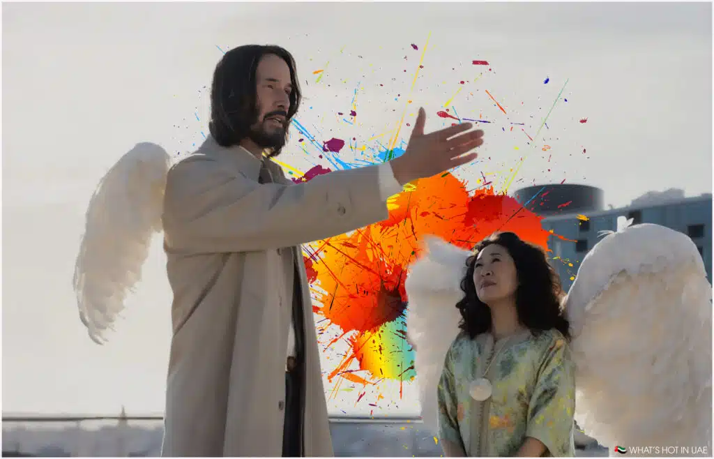 Two people wearing large white angel wings stand in front of a vibrant splash of multicolored paint, with one gesturing upwards.