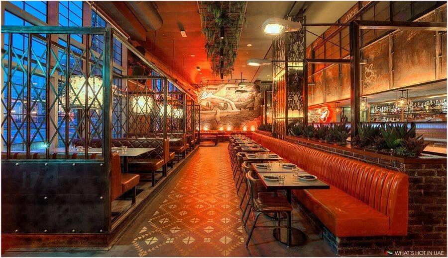 Interior of a stylish restaurant featuring red leather booth seating, industrial lighting, metal dividers, and a decorative patterned tile floor.