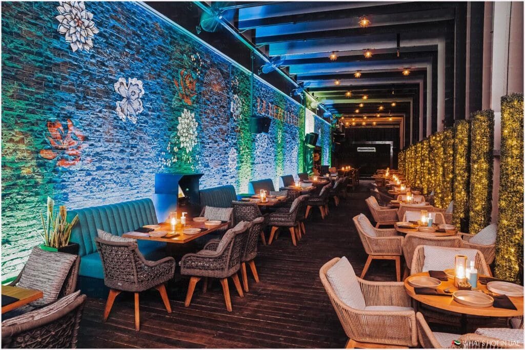 A stylish restaurant interior featuring a long corridor with wooden tables and woven chairs arranged along both sides. The left wall is decorated with illuminated flower designs, casting a colorful glow, while the right side is lined with greenery and string lights. Candles on each table enhance the warm, inviting atmosphere.