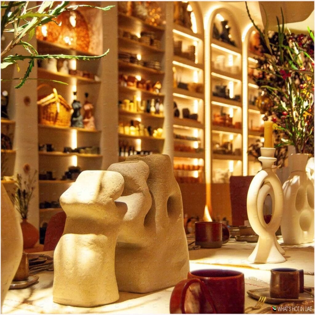 A warmly lit interior of a cafe or restaurant with a decorative table setup, featuring stone sculptures, round candlestick holders, coffee mugs, and shelves filled with pottery and other items in the background.