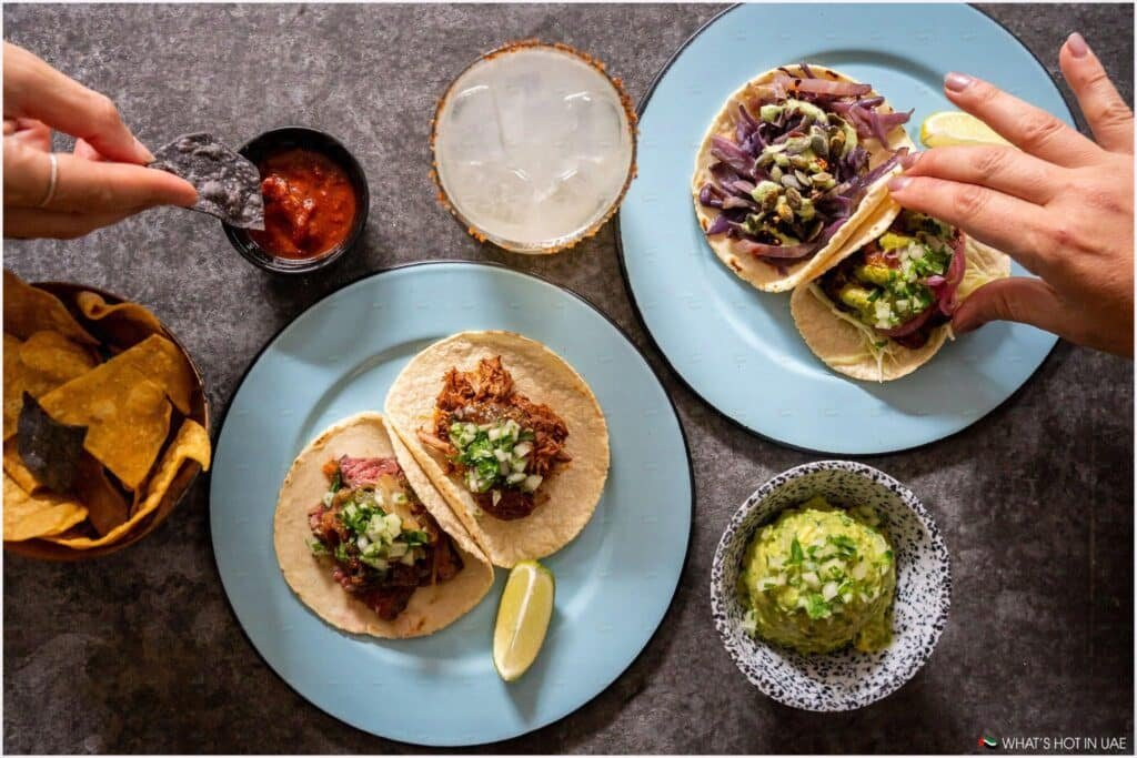 An overhead view of a table with two plates of assorted tacos, a bowl of salsa, a bowl of guacamole, a cocktail, and a basket of tortilla chips. A hand is dipping a blue corn chip into the salsa.