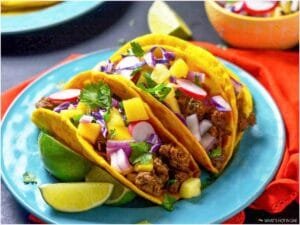 Three tacos filled with ground meat, diced mango, onions, radishes, and cilantro served on a turquoise plate with lime wedges.