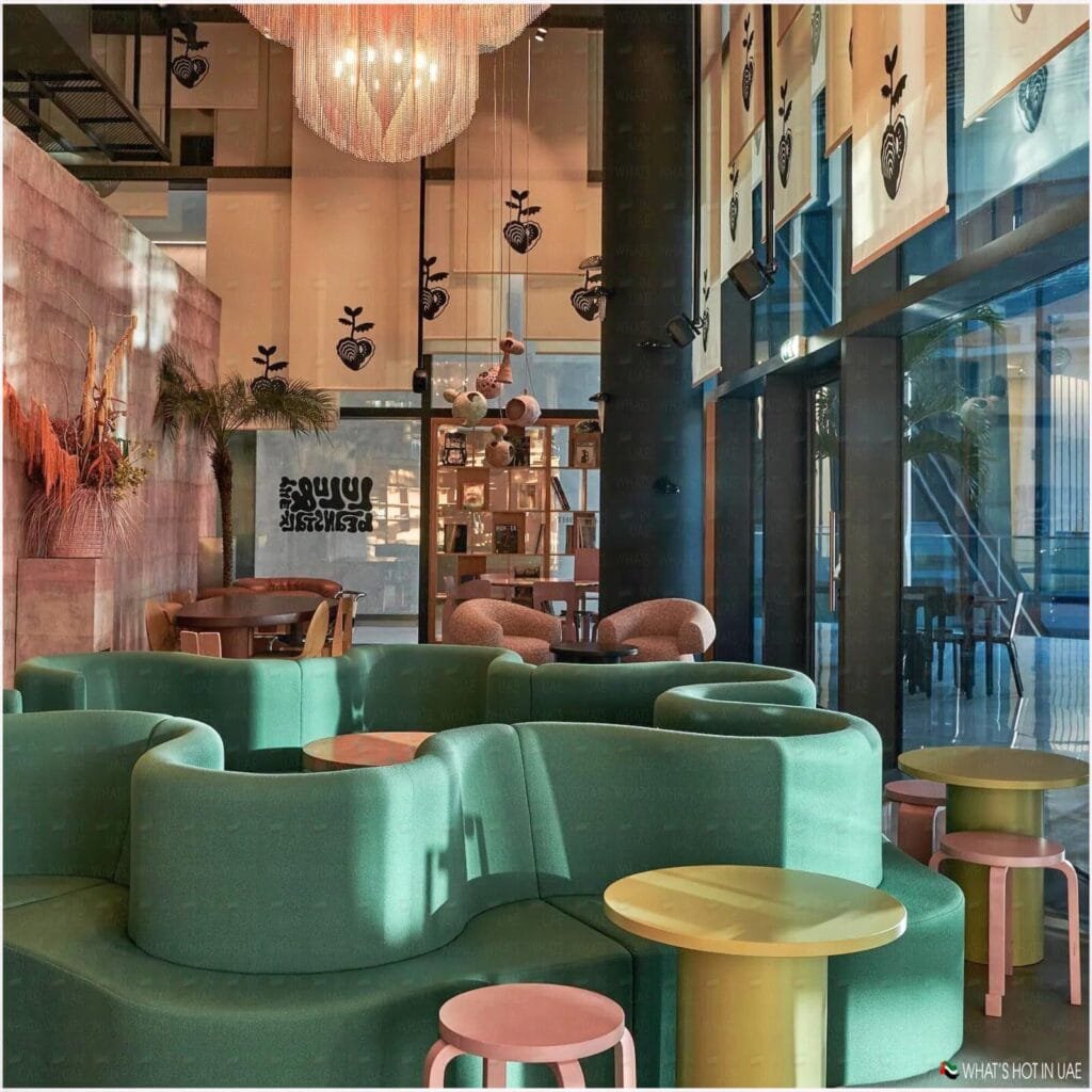 A modern cafe interior with green curved sofas, colorful round tables, and decorative lighting fixtures, featuring a large window allowing natural light to illuminate the space.