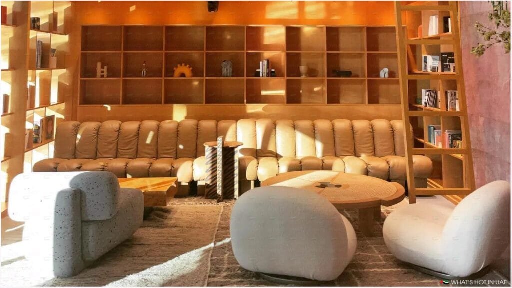 A cozy living room with a large tan leather sofa, two white armchairs, and a round wooden coffee table. The walls feature wooden shelves with decorative items and books, and a ladder for access. Soft sunlight streams through a window, creating a warm ambiance.