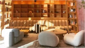 A cozy living room with a large tan leather sofa, two white armchairs, and a round wooden coffee table. The walls feature wooden shelves with decorative items and books, and a ladder for access. Soft sunlight streams through a window, creating a warm ambiance.