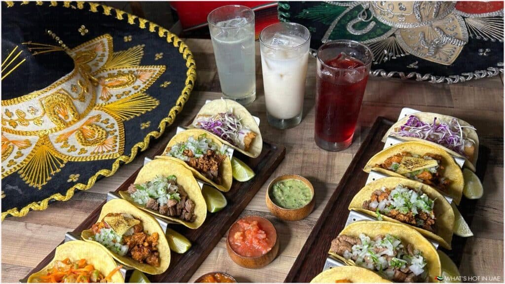 A colorful arrangement of three platters of tacos garnished with chopped onions and cilantro, accompanied by lime wedges, wooden bowls of salsa, and guacamole. Three drinks, including a light-colored beverage and a darker red one, are served alongside. Two ornate Mexican sombreros with golden and silver embroidery are also present on the table.