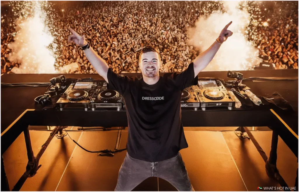 DJ Martin Garrix standing behind a mixing console on stage with arms raised, facing a massive crowd with fireworks in the background.