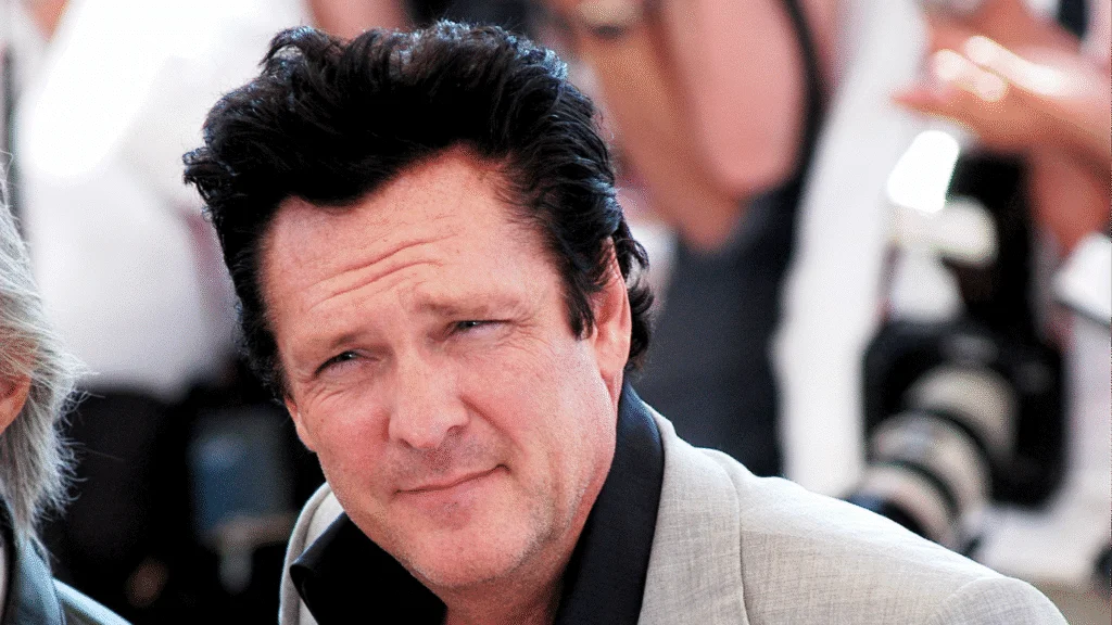 A man with short, dark, styled hair and a slight stubble is looking directly at the camera, wearing a light grey blazer over a black shirt, with soft-focus figures and cameras in the background.