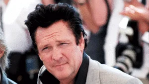 A man with short, dark, styled hair and a slight stubble is looking directly at the camera, wearing a light grey blazer over a black shirt, with soft-focus figures and cameras in the background.