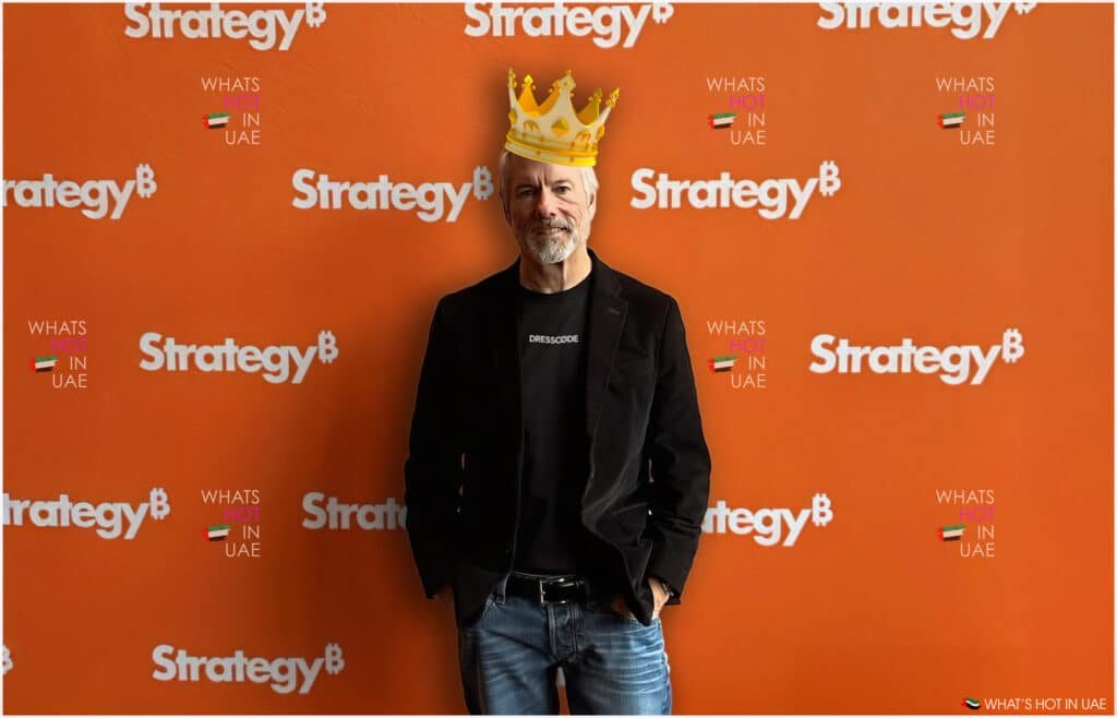 A person wearing a black jacket and jeans stands against an orange backdrop with 'Strategy' and 'What's Hot in UAE' logos, while a crown graphic is placed on their head. Michael Saylor to Speak at Bitcoin MENA