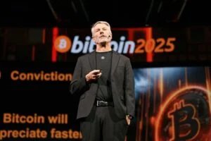 A person in black suit and t-shirt is speaking on stage at a Bitcoin 2025 conference, with a large Bitcoin logo and text in the background.