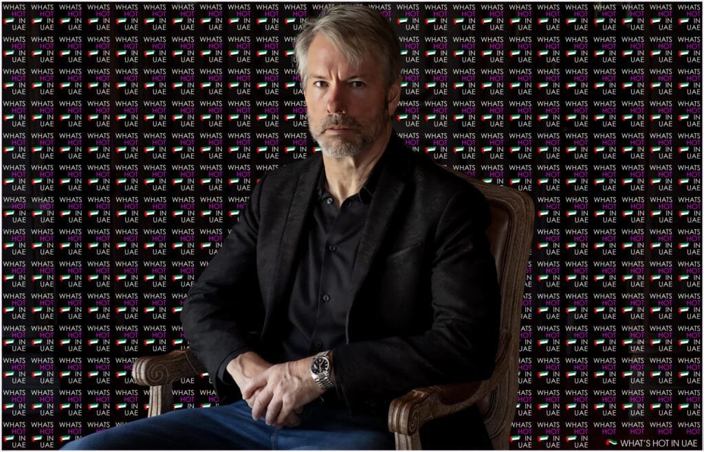 A man with gray hair and a beard is seated wearing a black jacket and a watch, against a backdrop featuring the repeated text "WHAT'S HOT IN UAE" and the UAE flag.