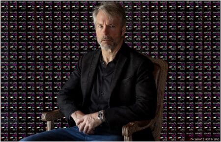 A man with gray hair and a beard is seated wearing a black jacket and a watch, against a backdrop featuring the repeated text "WHAT'S HOT IN UAE" and the UAE flag.