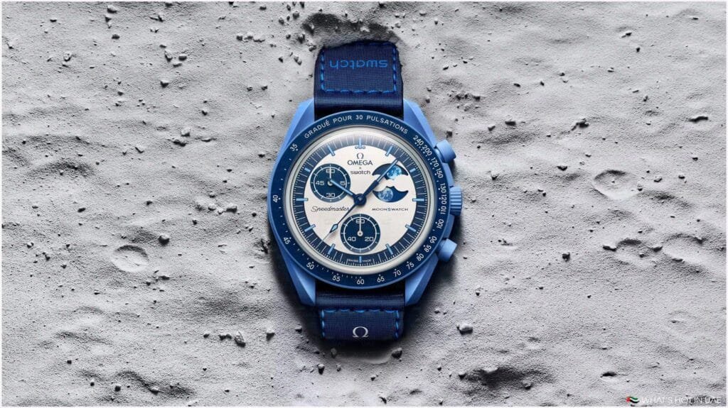 A blue Omega x Swatch Speedmaster Moonswatch resting on a textured lunar surface, featuring a white dial with three black sub-dials and a matching blue fabric strap.