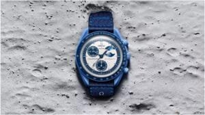 A blue Omega x Swatch Speedmaster Moonswatch resting on a textured lunar surface, featuring a white dial with three black sub-dials and a matching blue fabric strap.
