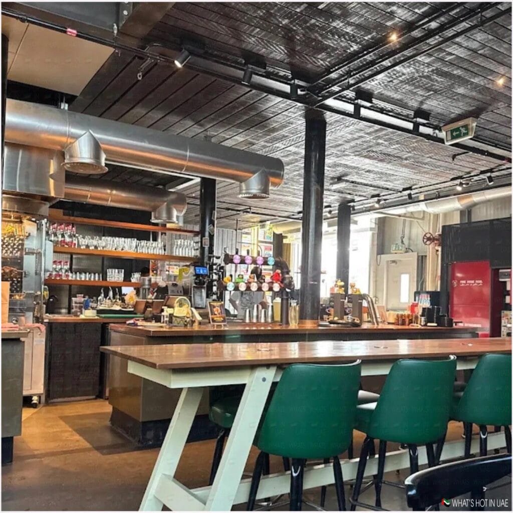 Interior of a modern bar with a long wooden table surrounded by green chairs, shelves stocked with glasses and bottles behind the bar, and industrial style metal ductwork on the ceiling.