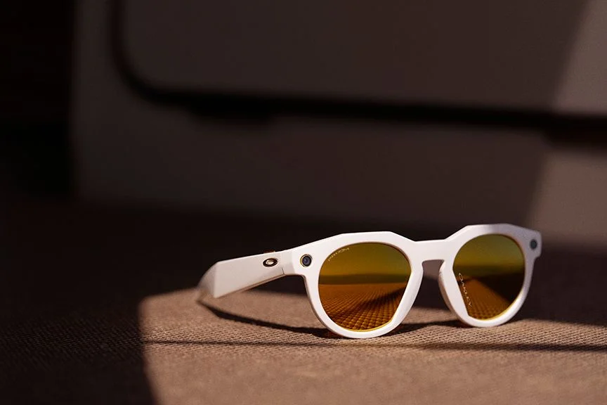 A pair of stylish white sunglasses with reflective yellow lenses resting on a textured surface, partially illuminated by light, creating a shadow.