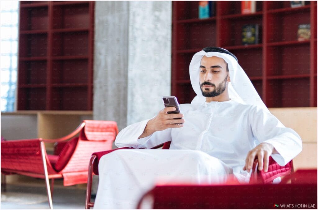 A man wearing traditional Middle Eastern attire is sitting on a red chair and looking at a smartphone. He is in a modern, well-lit room with bookshelves in the background.