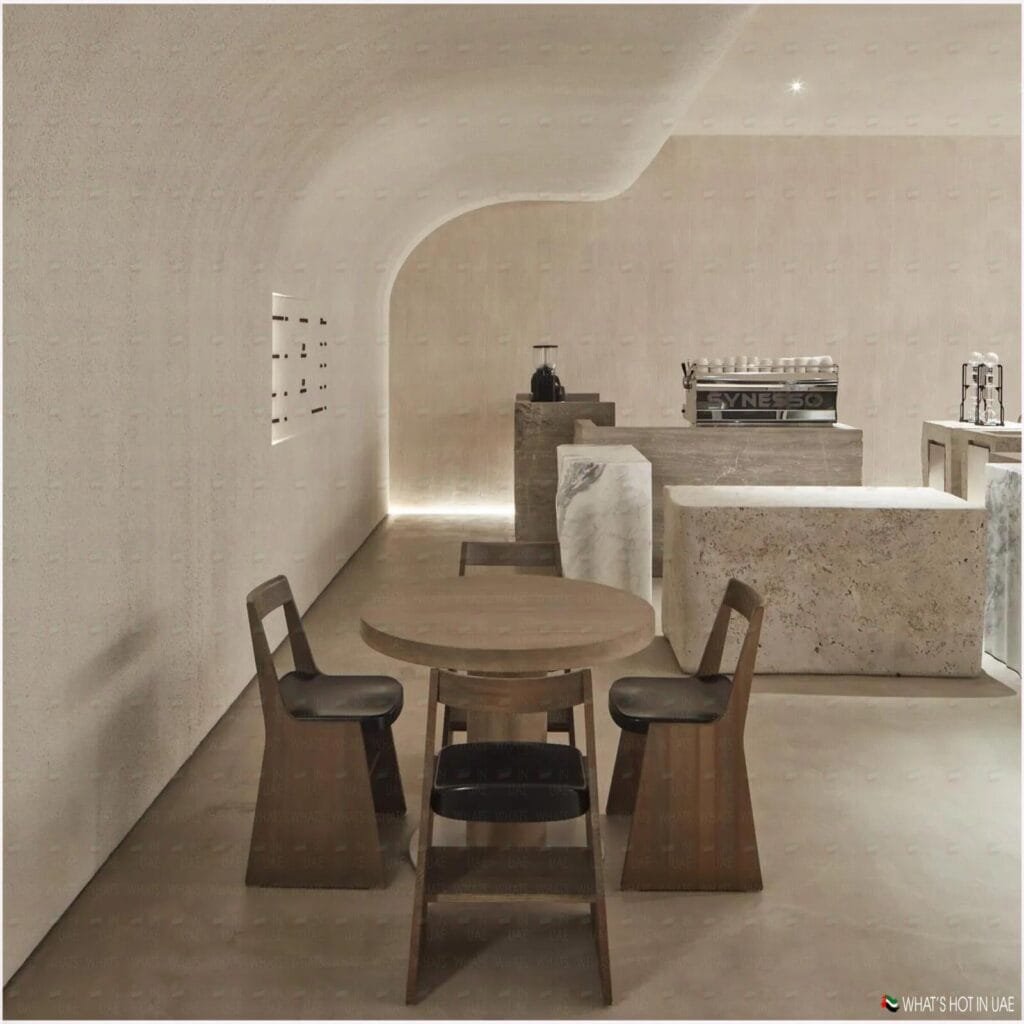 Minimalist cafe interior with a round wooden table and three wooden chairs in the foreground, a modern coffee machine on a marble counter in the background, and soft lighting accentuating the curved, textured walls.