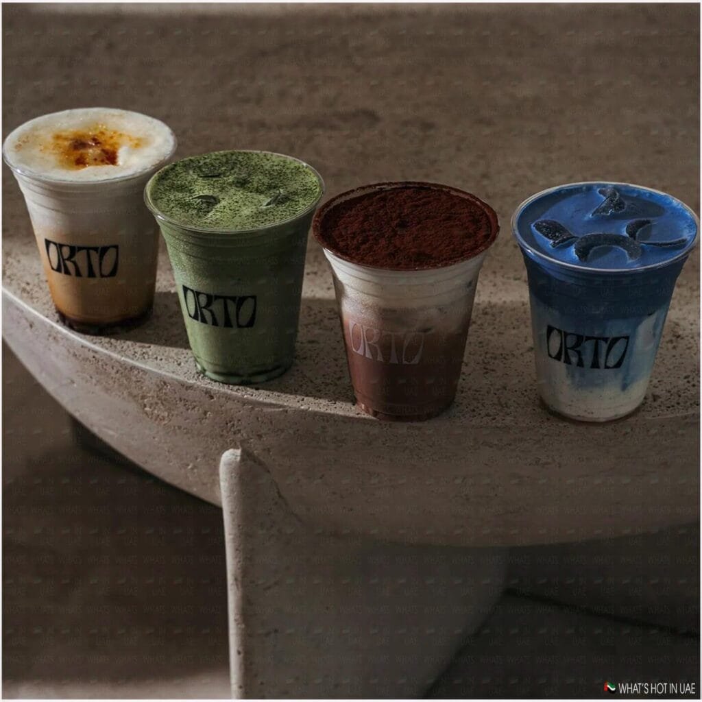 Four different flavored iced beverages in clear plastic cups are placed on a stone ledge, each labeled with 'ORTO'. The drinks include a caramel-topped latte, a matcha latte with green tea powder, a chocolate drink with a cocoa powder topping, and a vibrant blue beverage with three leaf-shaped ice cubes.