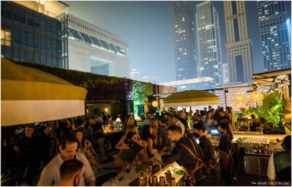 A bustling rooftop bar in an urban setting during the evening, with a crowd of people socializing and bartenders serving drinks. Tall skyscrapers are visible in the background, illuminated against the night sky.