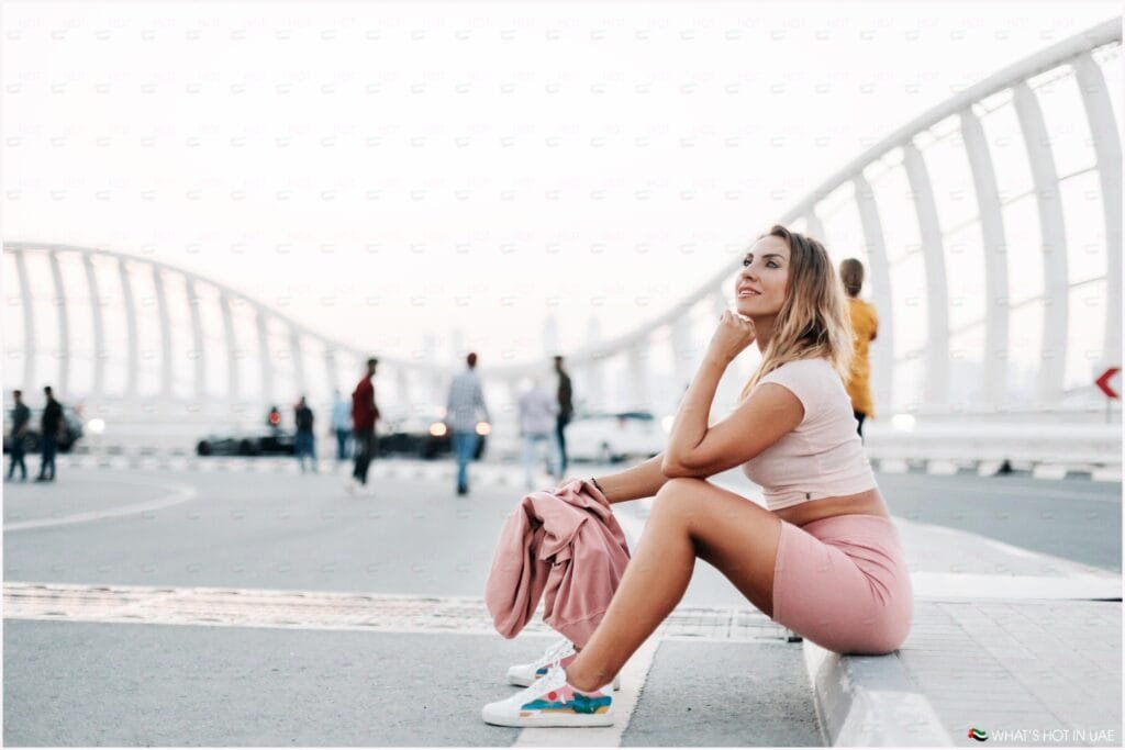 A woman in a pink outfit sitting on a ledge of a modern arched bridge, looking upwards with a thoughtful expression, while holding a pink jacket. The bridge has white, skeletal arches, with several pedestrians walking in the background.