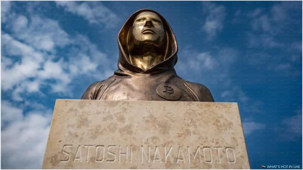 A bronze statue of a hooded figure representing Satoshi Nakamoto, with a Bitcoin symbol on its chest, set against a blue sky with clouds.
