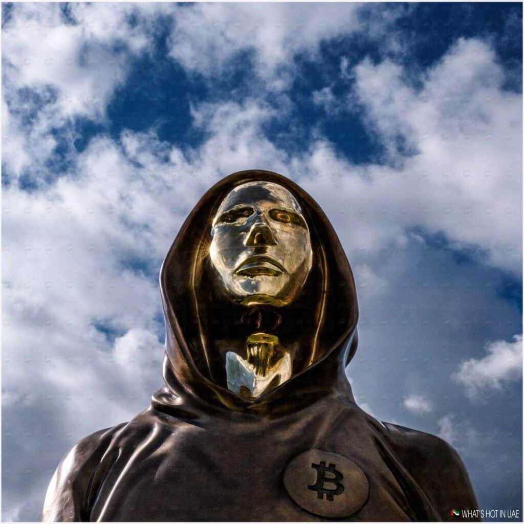 A bronze statue of an anonymous figure wearing a hooded robe with a reflective gold face, set against a partly cloudy sky. The robe features a Bitcoin symbol on the chest.